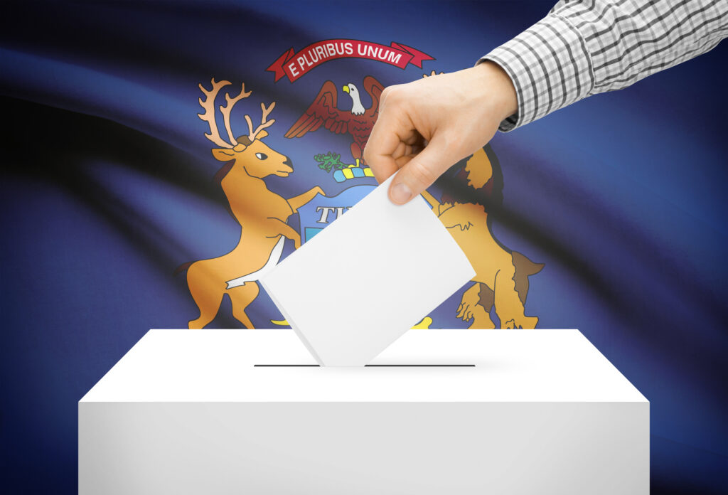 An image depicting a hand placing a ballot in a box during elections in front of the flag of the state of Michigan. The flag is blue with the state seal in the center.